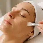 Ruka kozmetičarke nanosi enzimski piling na lice opuštene žene u mirnom spa ambijentu.