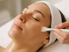 Ruka kozmetičarke nanosi enzimski piling na lice opuštene žene u mirnom spa ambijentu.