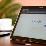 Tablet sa otvorenom Google početnom stranicom na stolu, uz šalicu kave, pametni telefon i olovku.